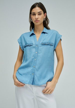 Femme portant une chemise en denim bleu clair sans manches avec boutons sur le devant et un pantalon blanc, debout devant un fond uni.