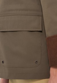 Καφέ υφασμάτινο σακάκι με μεγάλη τσέπη, υφασμάτινα καφέ κορδόνια με υφή στις μανσέτες και μεταλλικές λεπτομέρειες με κουμπιά κοντά στο τελείωμα.