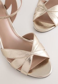 Sandali con tacco alto dorati con design a incrocio, texture metallica liscia, punta aperta e soletta imbottita. Dettagli suola nera.