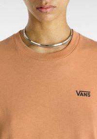 Maglietta Vans color marrone ruggine con colletto tondo e maniche corte, presenta un logo nero sul lato sinistro del petto. Tessuto di cotone liscio. Collana choker argentata.