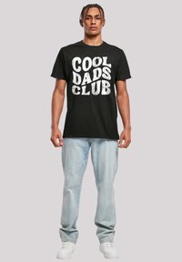 Zwarte katoenen T-shirt met gedurfde witte "COOL DADS CLUB" opdruk. Gecombineerd met lichtblauwe denim jeans en witte sneakers.