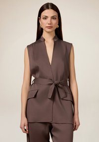 Bruin mouwloos vest met een mandarinnekraag, tailleband met strik en twee voorzakken, gemaakt van een gladde stof met een matte afwerking.