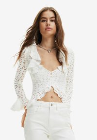Top bianco a maniche lunghe con scollatura arricciata, chiusura con bottoni e motivo ritagliato. Presenta maniche svolazzanti e un design corto. Abbinato a pantaloni bianchi.