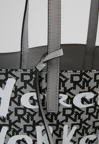 Gros plan d’un sac cabas noir et blanc avec un motif de lettres répétées et de grandes lettres en gras, avec des poignées et coutures en cuir gris clair.