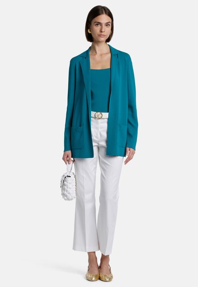 Blazer turquoise sur un top turquoise, associé à un pantalon blanc et des chaussures avec des accents dorés. Porte un sac à main blanc texturé avec des quincailleries dorées.