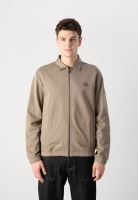Béžová bunda na zips s golierom, bočnými vreckami, rebrovými manžetami a malým logom na hrudi, skombinovaná s tmavými džínsami.
