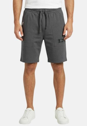 Homme portant un short gris foncé avec cordon de serrage, poches plaquées à l'avant, et des baskets blanches, debout devant un fond blanc.