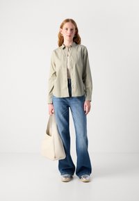 Light green button-up shirt over a beige top, paired with flared blue jeans and a cream shoulder bag. Sneakers with a metallic finish.