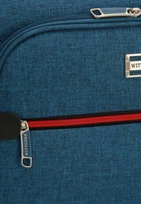 Sac en tissu bleu avec une fermeture éclair rouge, des accents noirs et un détail en métal au logo. La texture semble tissée, avec une finition lisse sur les accessoires.