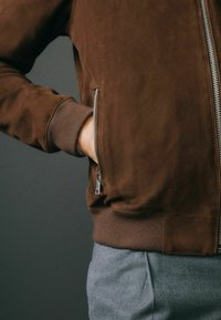 Chaqueta bomber de ante marrón con puños acanalados, que presenta un cierre de cremallera plateado y un bolsillo lateral, combinada con pantalones de color gris claro. Textura suave.
