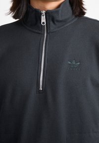 Pullover negro con cremallera hecho de tejido texturizado. Presenta un cuello alto y un logo de Adidas bordado en verde en el pecho.