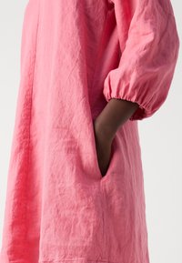 Vue de profil d'une personne portant une robe rose ample avec des manches bouffantes jusqu'au coude, main placée dans la poche latérale.