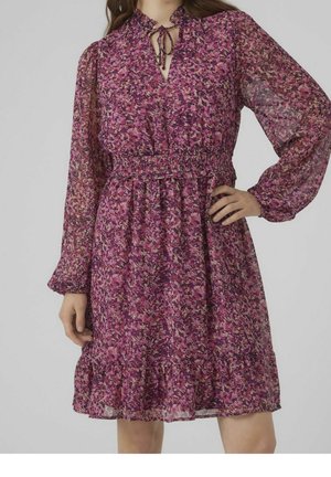 Vrouw die een paarse bloemenjurk met lange mouwen, een geplooide taille en een gerimpelde zoom draagt, staat met één hand op haar heup.
