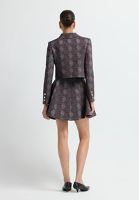 Mujer con el cabello recogido en un moño, vestida con un conjunto de blazer y falda a juego con estampado, y tacones de kitten negros, de pie frente a un fondo claro y liso.