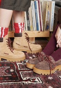 Botas de ante marrón y cuero marrón con cordones, suelas gruesas beige, y zapatos de ante púrpura y beige, ambos con cordones de diseño, sobre una alfombra texturizada.