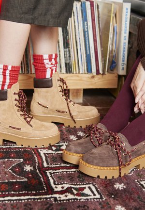 Des bottes en daim tan et en cuir marron à lacets avec des semelles chunky beige, et des chaussures en daim violet et beige, toutes deux avec des lacets à motifs, sur un tapis texturé.