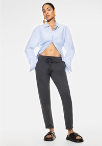 Lichtblauw overhemd met knoopsluiting, losse pasvorm met opgerolde mouwen, grijze joggers met trekkoord in de taille, en zwarte slidesandalen met dikke zolen.