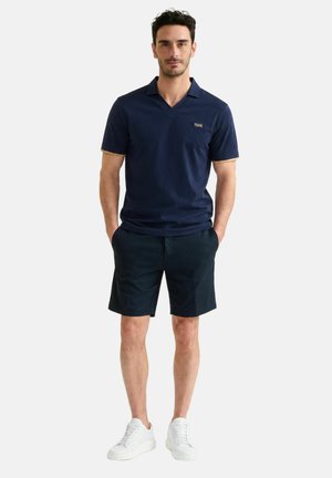 Uomo in piedi che indossa una polo blu navy a maniche corte, pantaloncini scuri e scarpe da ginnastica bianche, mani in tasca, su uno sfondo semplice e chiaro.