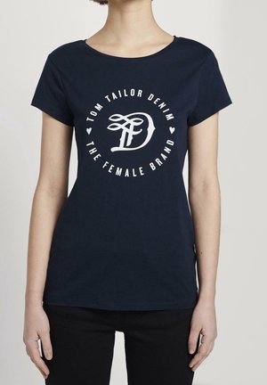 Kobieta ubrana w granatowy T-shirt z białym okrągłym napisem "Tom Tailor Denim, The Female Brand" oraz stylizowanym logo "D" na środku.