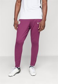 Burgundi sportnadrág, slim fit szabással, bal combközépén emblémával és kék kiegészítőkkel az oldalon. Fehér focicipővel viselve.
