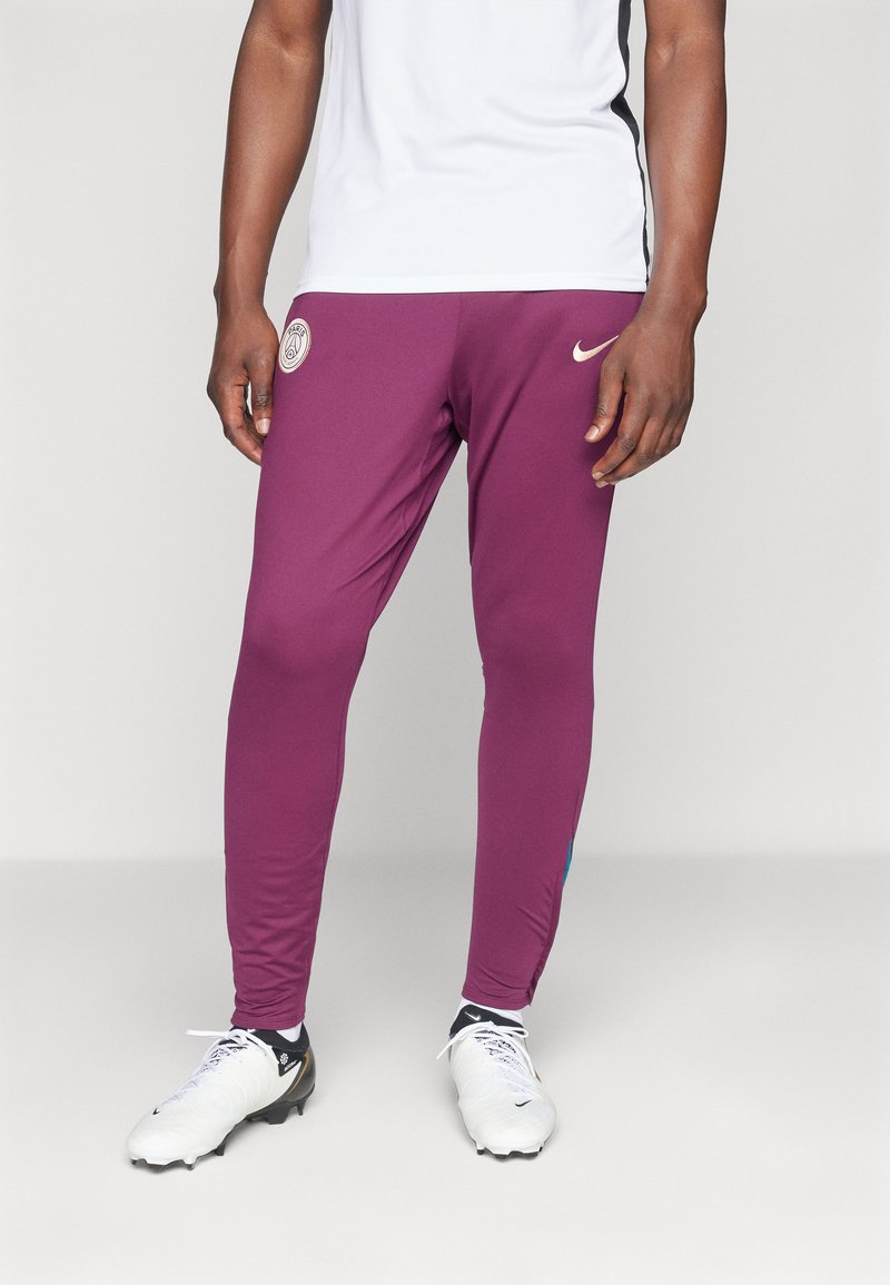 Burgundi sportnadrág, slim fit szabással, bal combközépén emblémával és kék kiegészítőkkel az oldalon. Fehér focicipővel viselve.