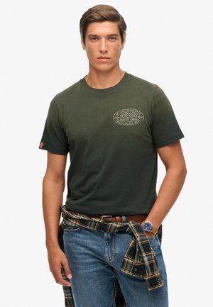 Mørkegrøn bomulds t-shirt med rund hals og et logo på brystet. Sammen med blå jeans og en ternet flannel skjorte bundet om taljen.