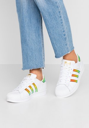 Stopy w białych butach sportowych Adidas Superstar z zielonymi, żółtymi i pomarańczowymi paskami oraz jasnoniebieskich krótkich dżinsach.