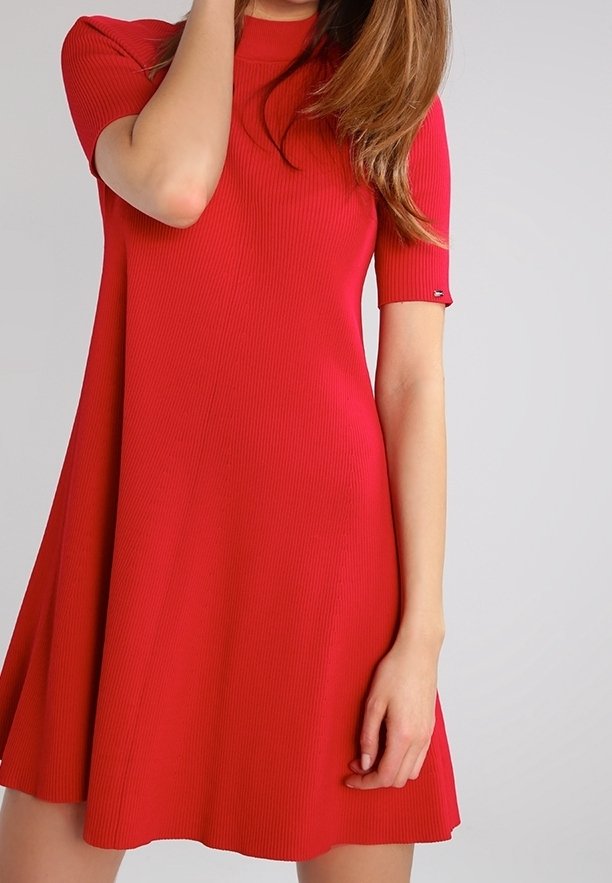 Femme portant une robe rouge à manches courtes, longueur genou, avec une texture côtelée, debout devant un fond gris uni.