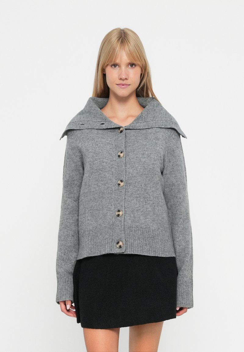 Pull en tricot gris avec un large col, fermeture par bouton et poignets côtelés, porté sur une jupe noire. Texture douce et poids moyen.