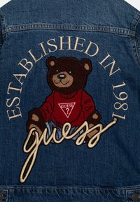 Jeans jakna s izvezenim medvjedom koji nosi crvenu majicu. "Osnovano 1981." i "guess" su ušivene u kontrastnim bojama.