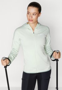 Lys grøn zip-up hettejakke med teksturert stoff, utstyrt med en frontlomme og brodert logo, sammen med svarte friluftsbukser og turstaver.