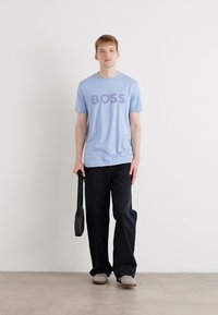 T-shirt bleu clair à col rond avec le texte 'BOSS', associé à un pantalon large noir et des chaussures gris à enfiler ; portant un sac à épaule noir.