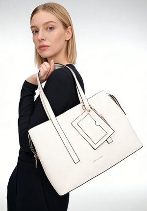 Jeune femme en noir tient un grand sac à main blanc avec double anse et porte-cartes attaché sur fond uni.