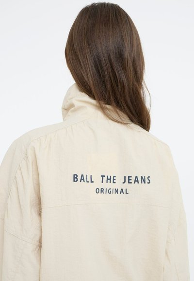 Veste oversize beige clair avec un col haut, comportant un texte bleu foncé dans le dos qui indique "BALL THE JEANS ORIGINAL." Texture lisse.