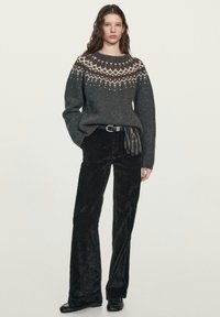 Femme portant un pull en tricot à motif foncé, pantalon en velours noir, ceinture noire, tenant des gants en cuir noir, debout devant un fond uni.