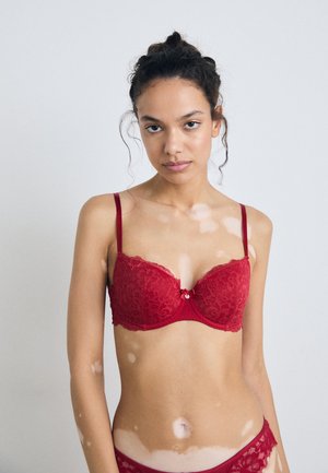 Soutien-gorge push-up en dentelle rouge avec des bretelles réglables et un détail en nœud au centre. Présente des bords en dentelle festonnés et une bande inférieure lisse.
