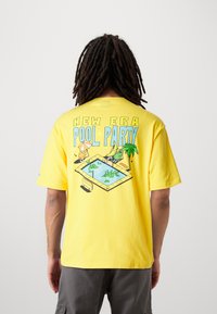 Camiseta de algodón amarilla con un diseño gráfico de una escena de fiesta en la piscina en la parte de atrás, con personajes y elementos tropicales. Mangas cortas.