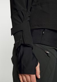 Czarna techniczna kurtka z marszczonymi rękawami, kieszenią na zamek oraz kontrastowymi materiałami dla lepszej tekstury i funkcji.