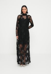 Rochie maxi de dantelă neagră, cu mâneci lungi și modele florale. Textură transparentă, decolteu înalt și design stratificat. Purtați cu pantofi de culoare deschisă.