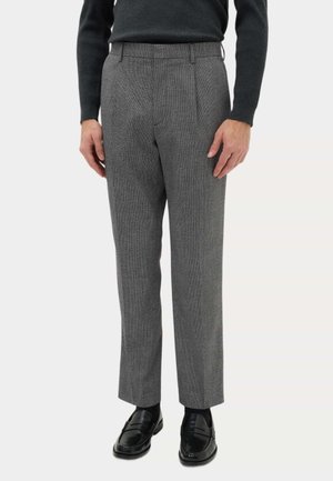 Mężczyzna w szarych teksturowanych eleganckich spodniach, czarnych skarpetkach, czarnych skórzanych loafersach i czarnym swetrze z długim rękawem, stojący z rozluźnionymi rękami.