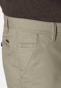 Beige Hosen mit strukturiertem Finish, ausgestattet mit einem roten Akzent am Bund und vier Taschen, einschließlich einer kleinen Münztasche.