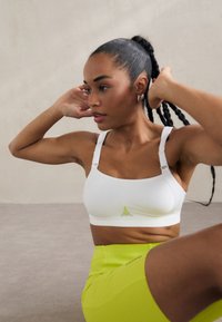 Sutien sport alb cu bretele late, o textură neted�ă și un mic logo galben accentuat la centru. Modelul poartă o piesă de jos verde strălucitor.