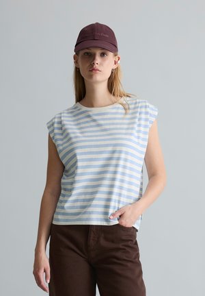 Jeune femme portant une casquette violette, un débardeur blanc à rayures bleu clair et un pantalon marron foncé, debout avec une main dans la poche.