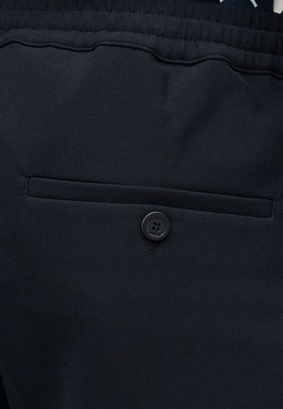 Gros plan sur la poche d'un pantalon bleu marine foncé avec une fermeture à bouton et un tissu texturé sous une ceinture élastique.