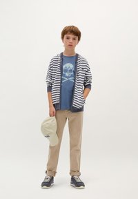 Rayas de sudadera en azul marino y blanco, camiseta azul con gráfico de calavera, pantalones beige, sosteniendo una gorra beige claro y vistiendo zapatillas de deporte azul marino.