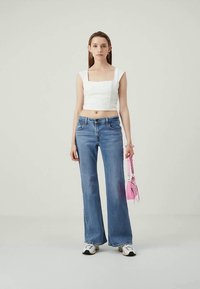 Giovane donna con top bianco corto, jeans a gamba larga blu, sneakers bianche, che tiene in mano una piccola borsa rosa, in piedi davanti a uno sfondo chiaro e neutro.