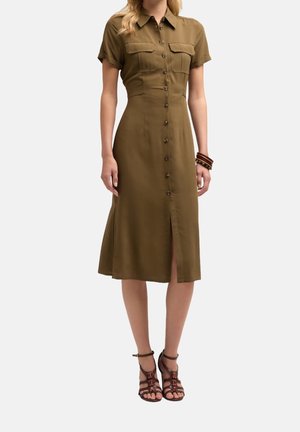 Kobieta ubrana w oliwkową sukienkę na guziki o długości do kolan, z krótkimi rękawami, przednimi kieszeniami oraz brązowymi, paskowanymi butami na obcasie z paskami wokół kostki.