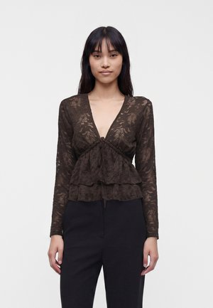 Femme portant un chemisier en dentelle marron foncé à manches longues avec un décolleté en V profond et un pantalon noir, debout devant un fond clair uni.