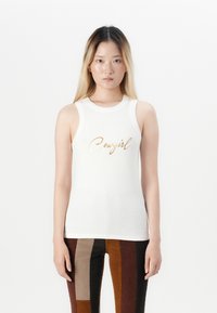 Hvit ermeløs tanktopp med grafikk av ordet «Cowgirl» i gullfolie, kombinert med brun- og rustfargede, vertikalt stripete smale bukser.