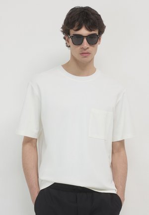 Un jeune homme avec des cheveux ondulés porte des lunettes de soleil noires, un t-shirt blanc à manches courtes avec une poche poitrine, et un pantalon noir devant un fond uni.